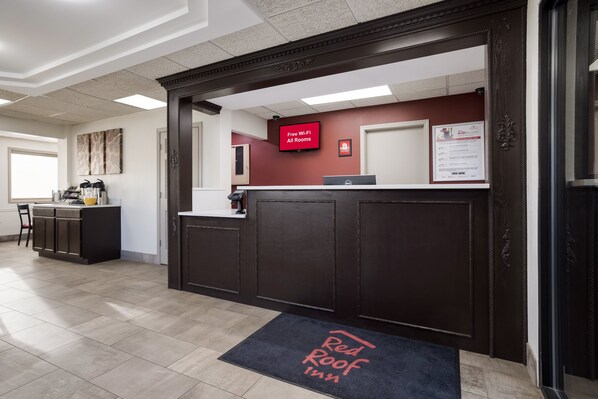 A reception area with a dark wood reception desk, a red wall-mounted sign indicating free Wi-Fi, and a digital screen. - Red Roof Inn Weedsport (Weedsport)