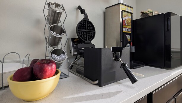 A kitchen counter with a waffle maker, a bowl of apples, and a waffle iron. - Red Roof Inn Weedsport (Weedsport)