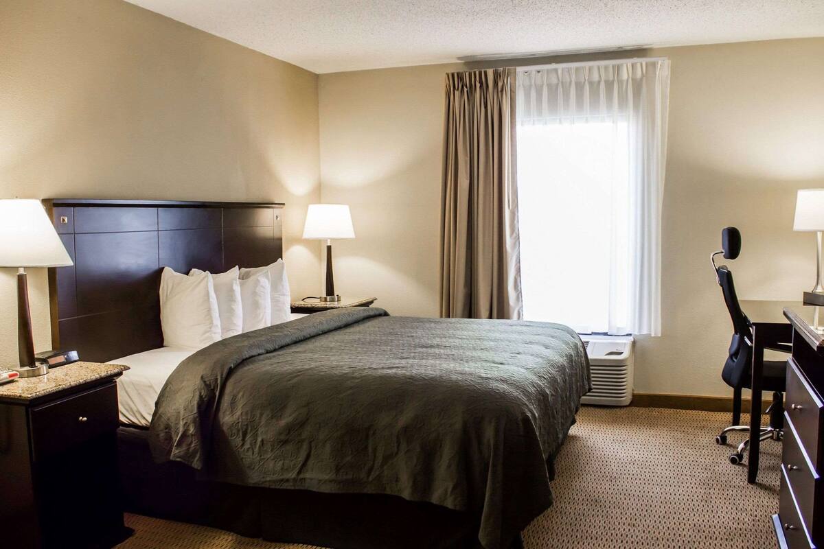 A hotel room with a large bed, two bedside tables with lamps, a desk with a chair, and a window with curtains.