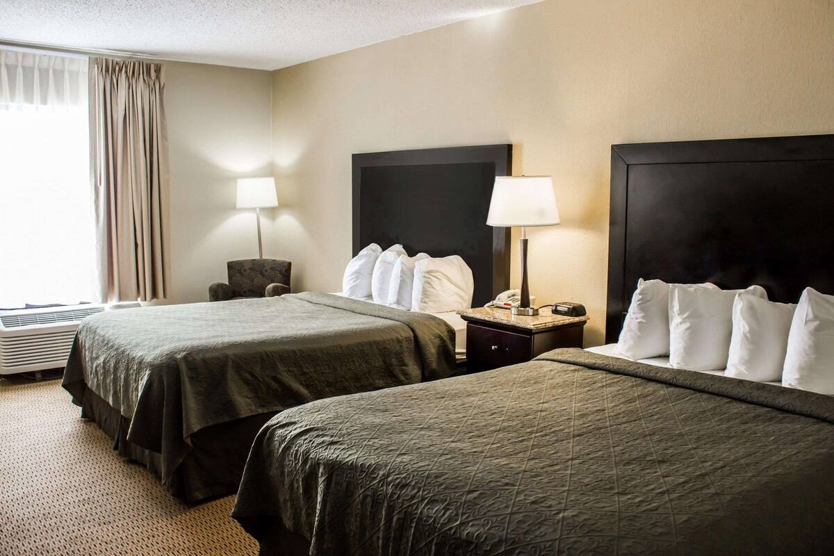 A hotel room with two beds, a chair, a desk with a lamp, and a large window with curtains.