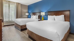 Quarto de hotel com duas camas, uma janela com cortinas, uma mesa de cabeceira com abajur e uma parede azul.