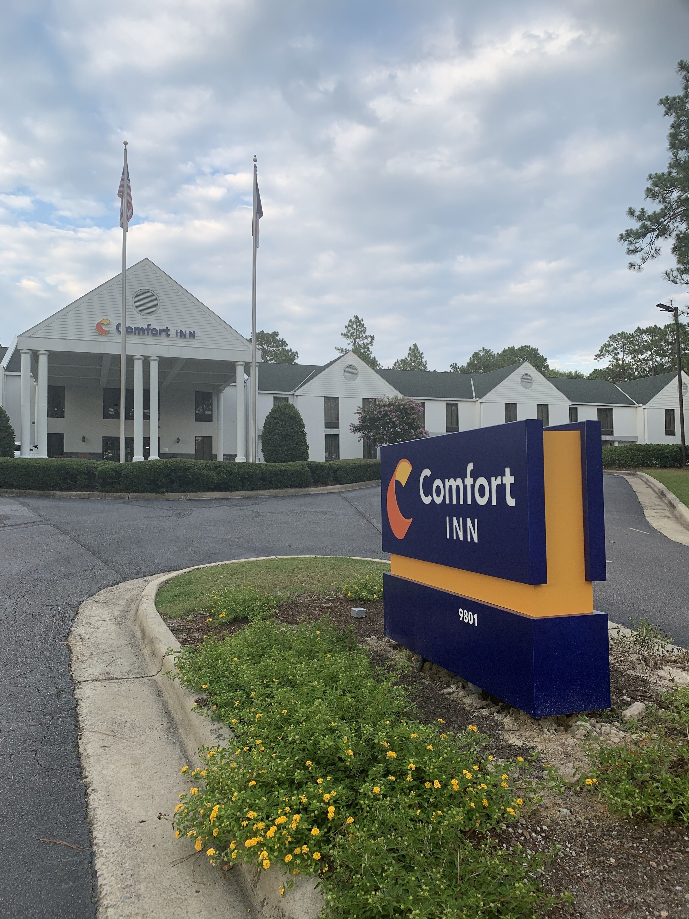 Un hôtel Comfort Inn, reconnaissable à son enseigne bien visible, à son mât de drapeau et à sa pelouse bien entretenue, agrémentée de fleurs jaunes.