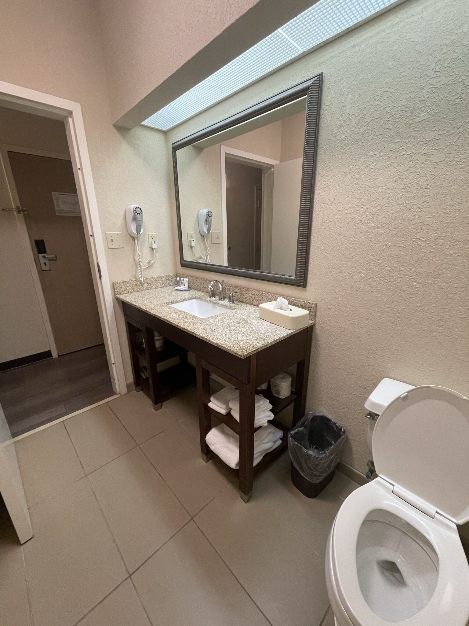 A bathroom with a double sink, a mirror, a hairdryer, a toilet, and a trash can.