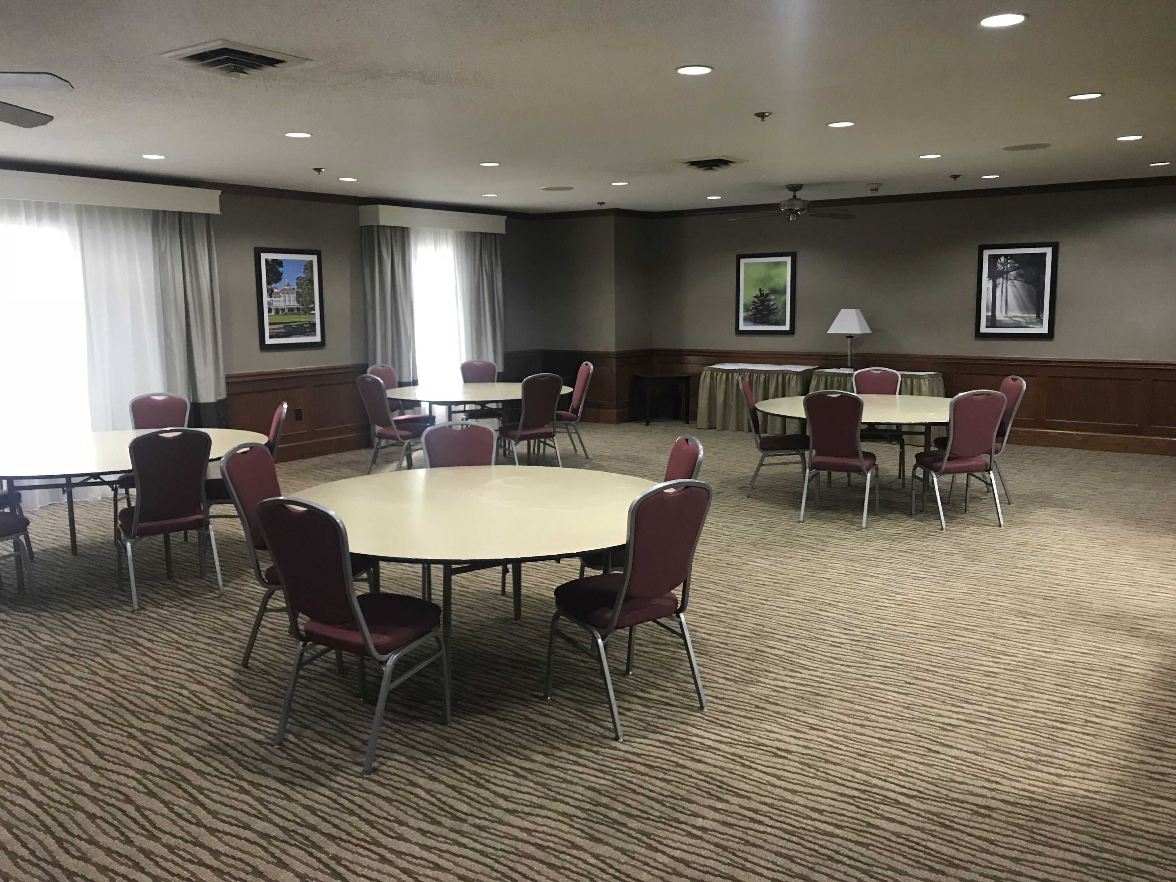Un salón de conferencias con mesas y sillas redondas, ventanas amplias con cortinas y cuadros en las paredes.