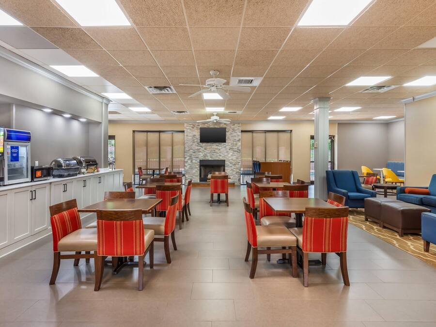 A hotel lobby with multiple dining tables and chairs, a fireplace, a television mounted on a stone wall, and a seating area with sofas and armchairs.