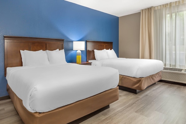 Habitación de hotel con dos camas, una mesita de noche, una lámpara y un ventanal con cortinas.