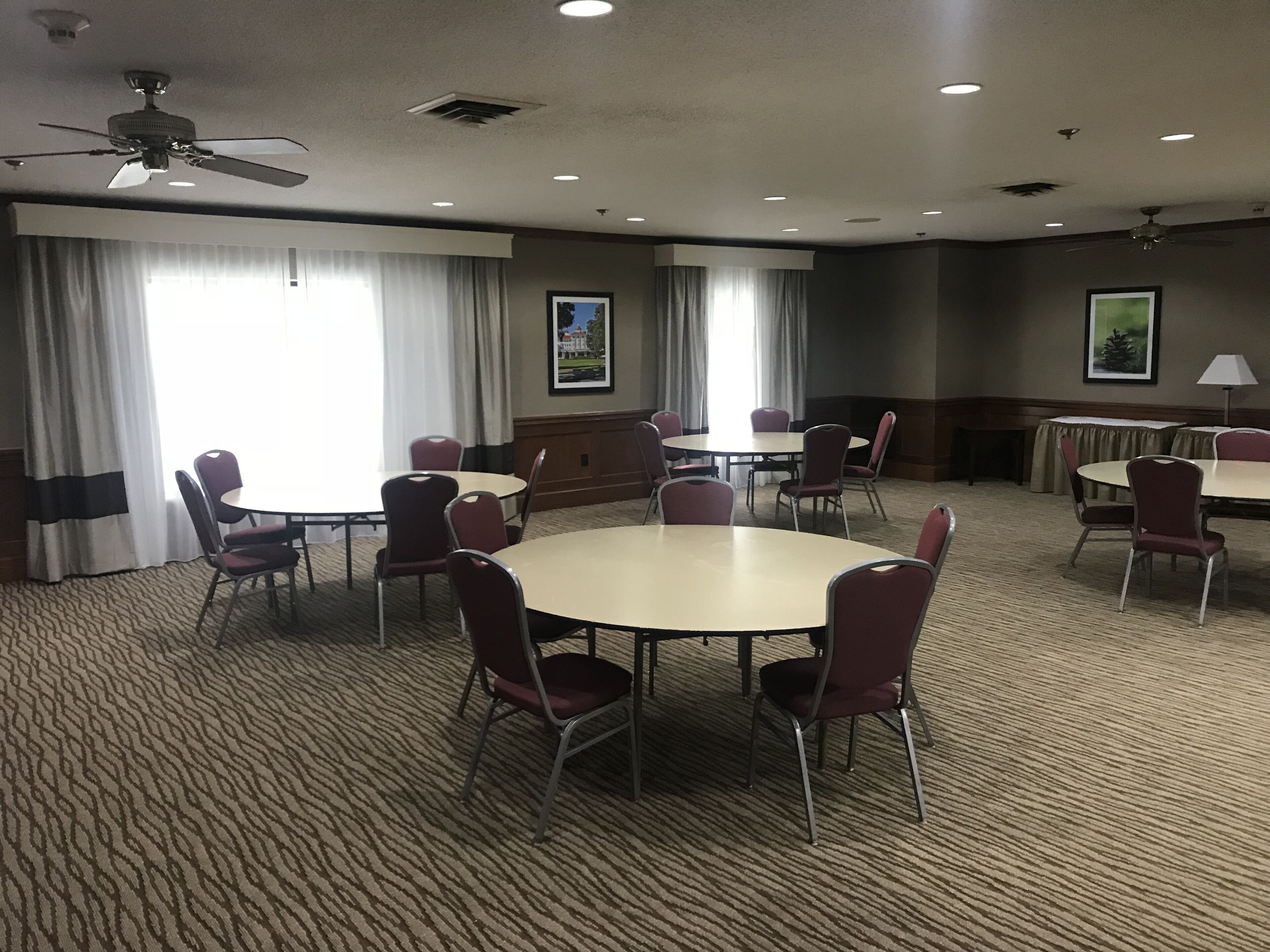 Un salón de conferencias con mesas y sillas redondas, ventanas amplias con cortinas y ventiladores de techo.