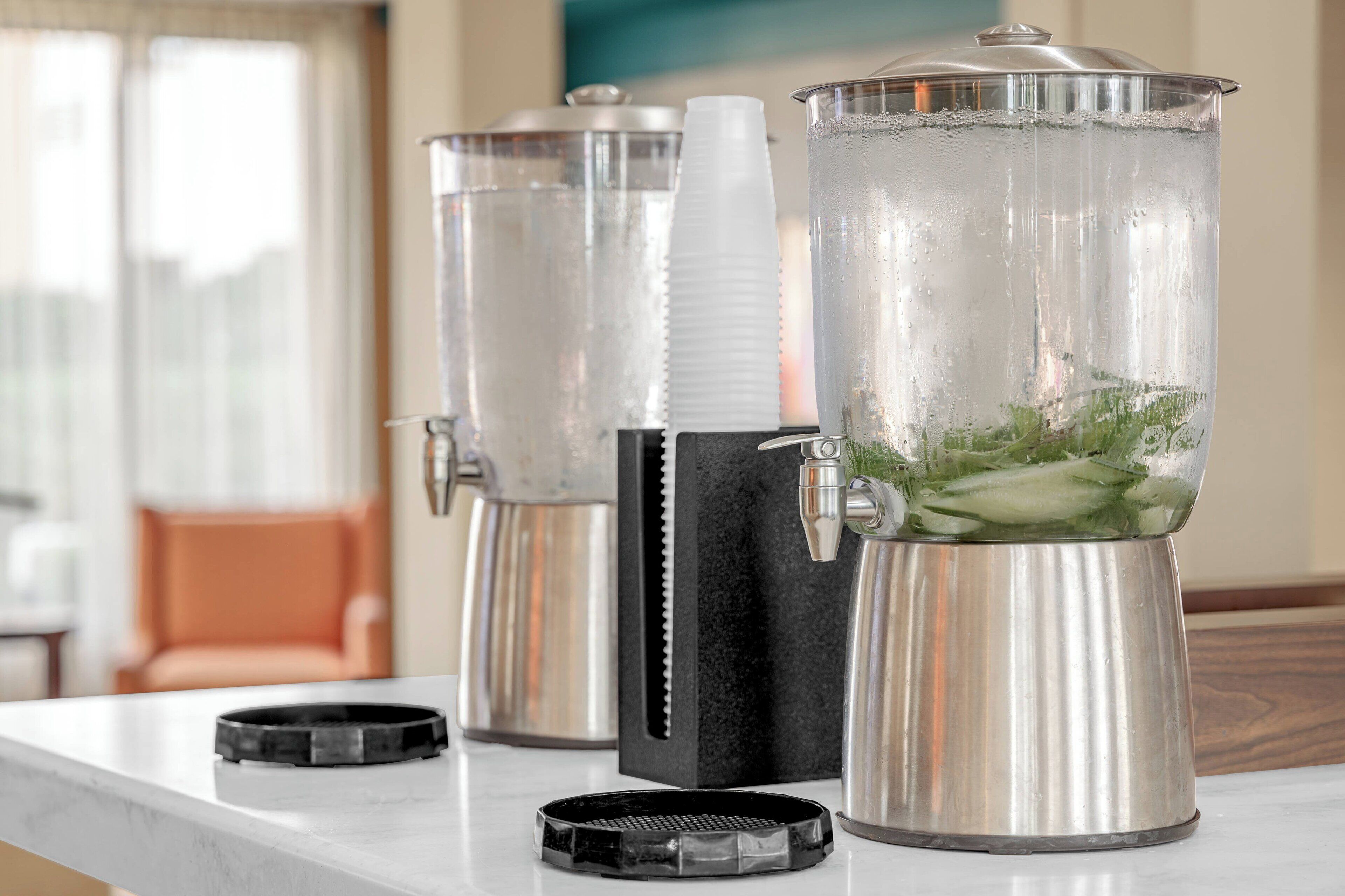 Tres dispensadores de bebidas de acero inoxidable sobre una encimera de mármol, con base negra y un recipiente transparente lleno de hielo y guarnición verde.