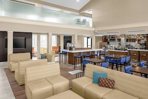 Un amplio vestíbulo de hotel con techos altos, una zona de estar con sofás y sillones, un área de bar con mostrador y taburetes, y una cocina visible al fondo.