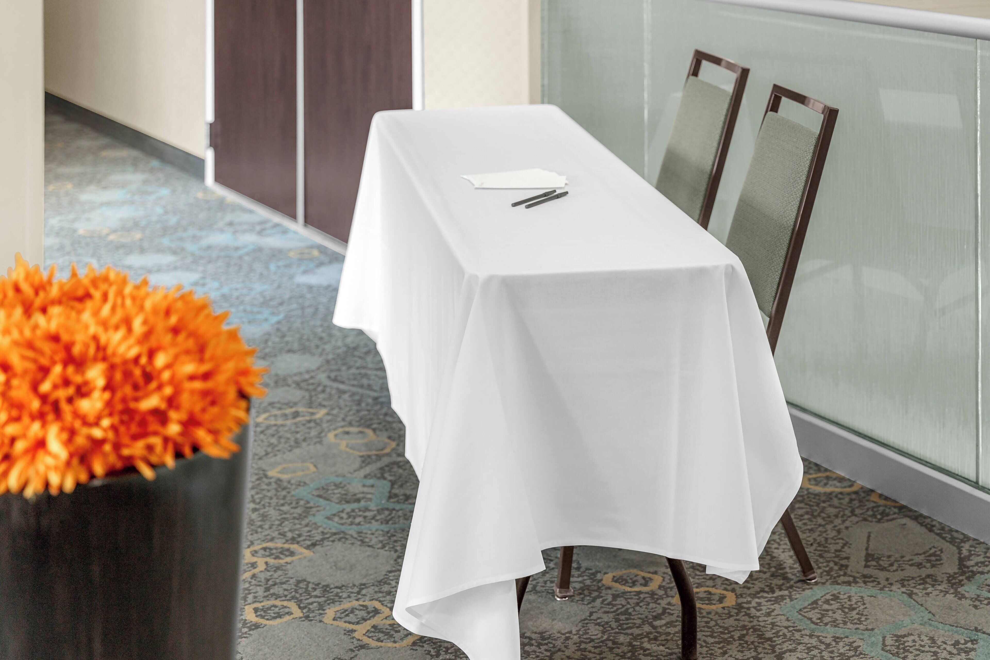 Et hotelværelse med et langt, hvidt bord, en vase med orange blomster og et mønstret tæppe.
