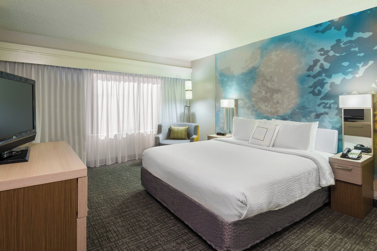 Habitación de hotel con una cama grande, un escritorio con televisor, una silla y una mesita de noche con lámpara.