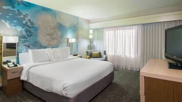 Quarto de hotel com uma cama grande, uma escrivaninha com televisão, uma cadeira, uma mesa de cabeceira com abajur e uma parede com desenho abstrato azul.