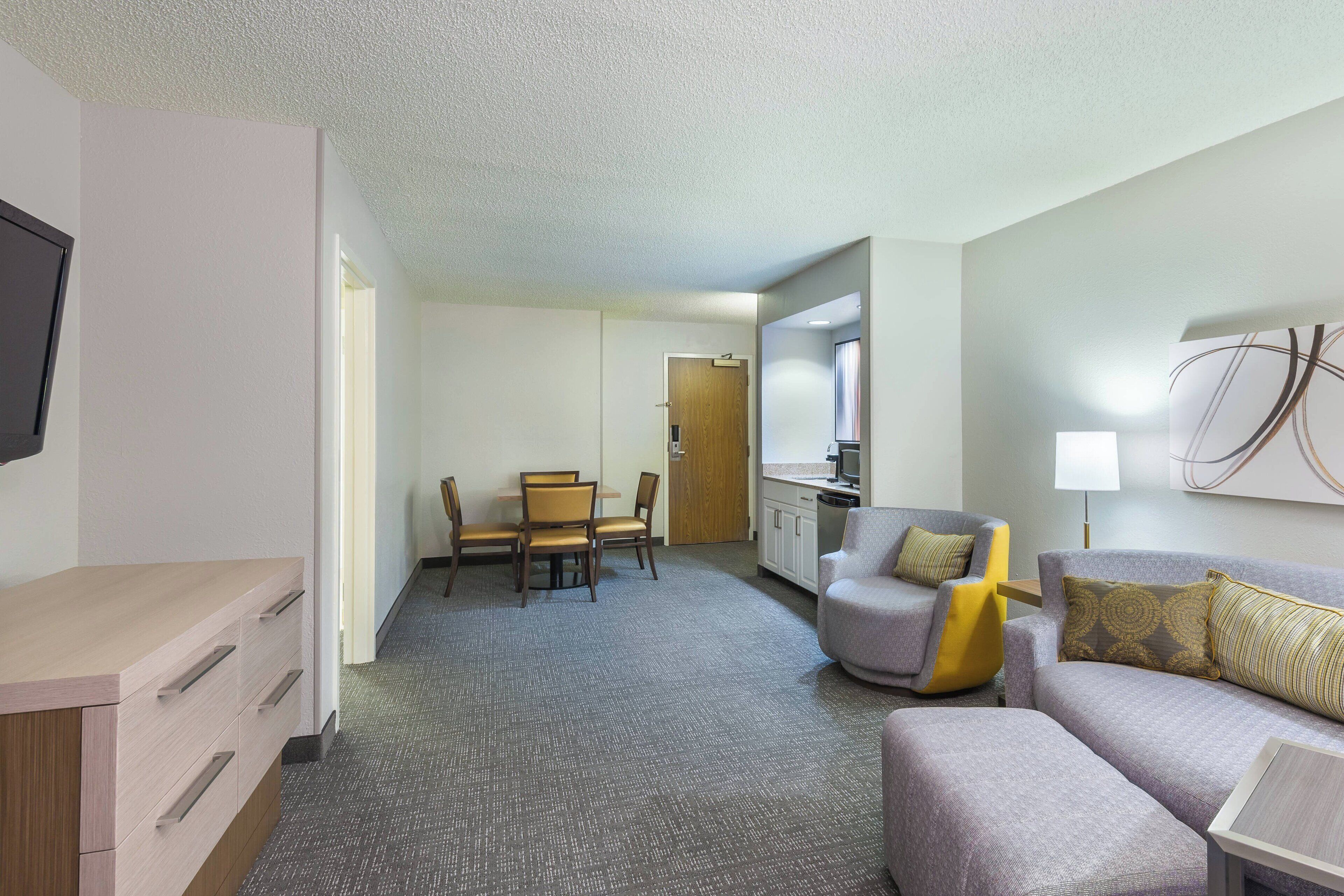 A hotel room with a sofa, a small table, a TV, a desk with drawers, a dining area with chairs, and a kitchen area.