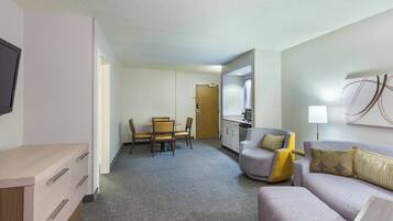 A hotel room with a sofa, a small table, a TV, a desk with drawers, a dining area with chairs, and a kitchen area.