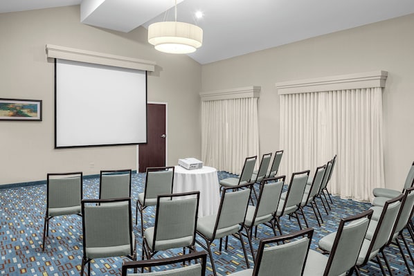 Un salón de conferencias con una pantalla de proyector, una mesa blanca y filas de sillas.