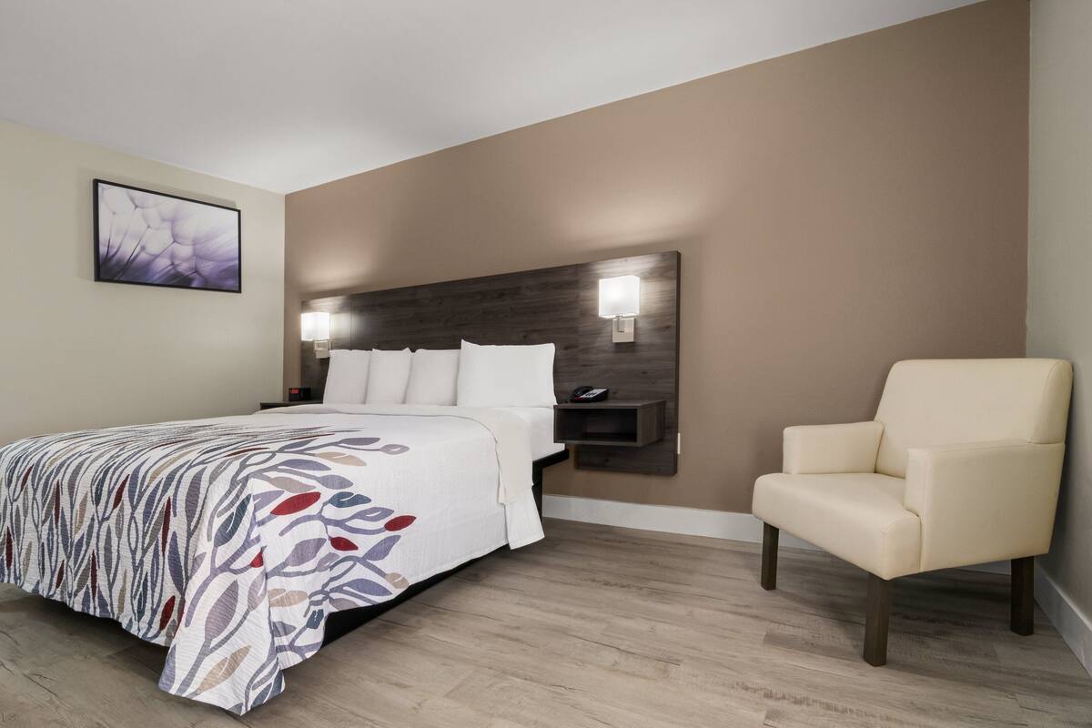 A hotel room with a bed, a chair, a nightstand, and a wall-mounted lamp.