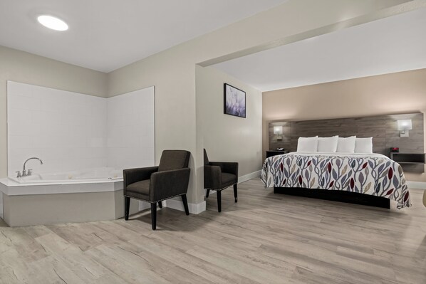 A modern bedroom with a large bed, a sitting area with two chairs, a bathroom with a bathtub, and a ceiling light.