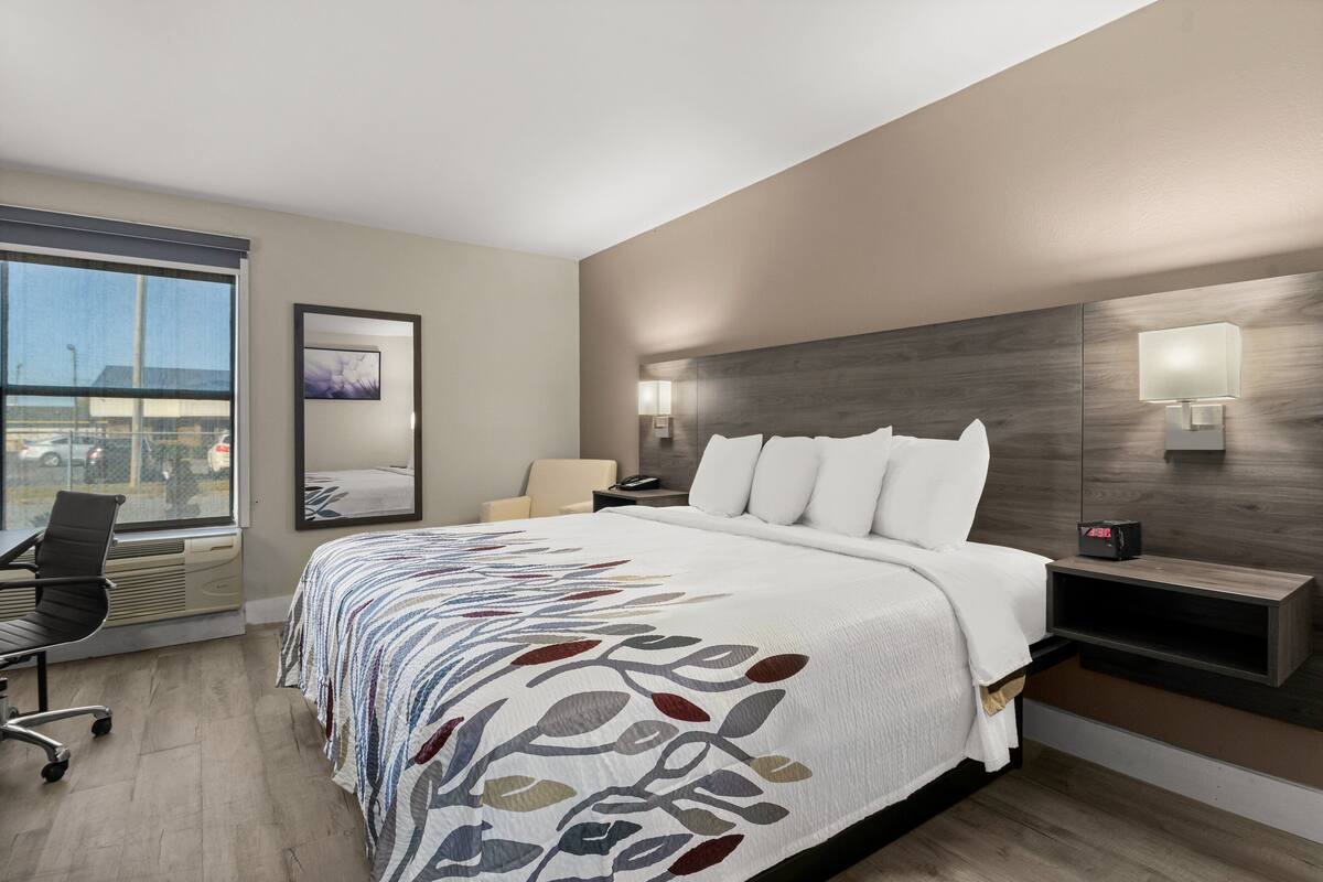 A hotel room with a large bed, a desk, a chair, a window with a view, and a wall-mounted lamp.