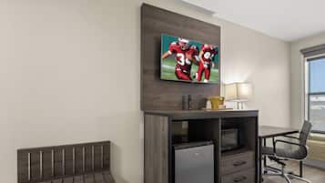 A hotel room with a desk, a small refrigerator, a bench, a television mounted on the wall, and a window with blinds.