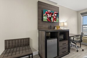A hotel room with a desk, a small refrigerator, a bench, a television mounted on the wall, and a window with blinds.