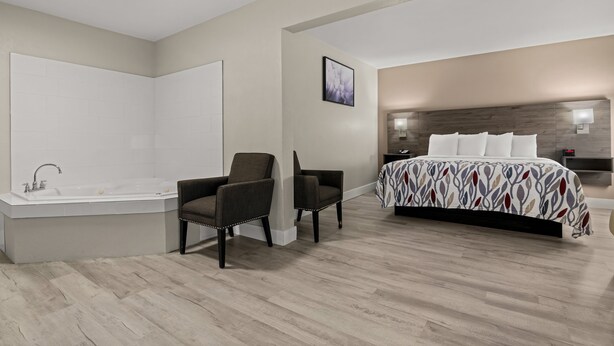 A modern bedroom with a large bed, a sitting area with two chairs, a bathtub, and a headboard with built-in lighting. - Red Roof Inn Washington, NC (Washington)