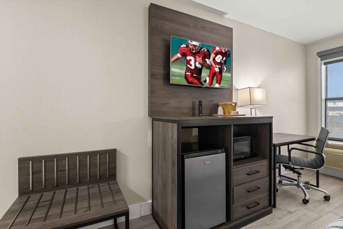 A hotel room with a wooden bench, a desk with a computer, a refrigerator, a TV mounted on the wall, and a window with a view.