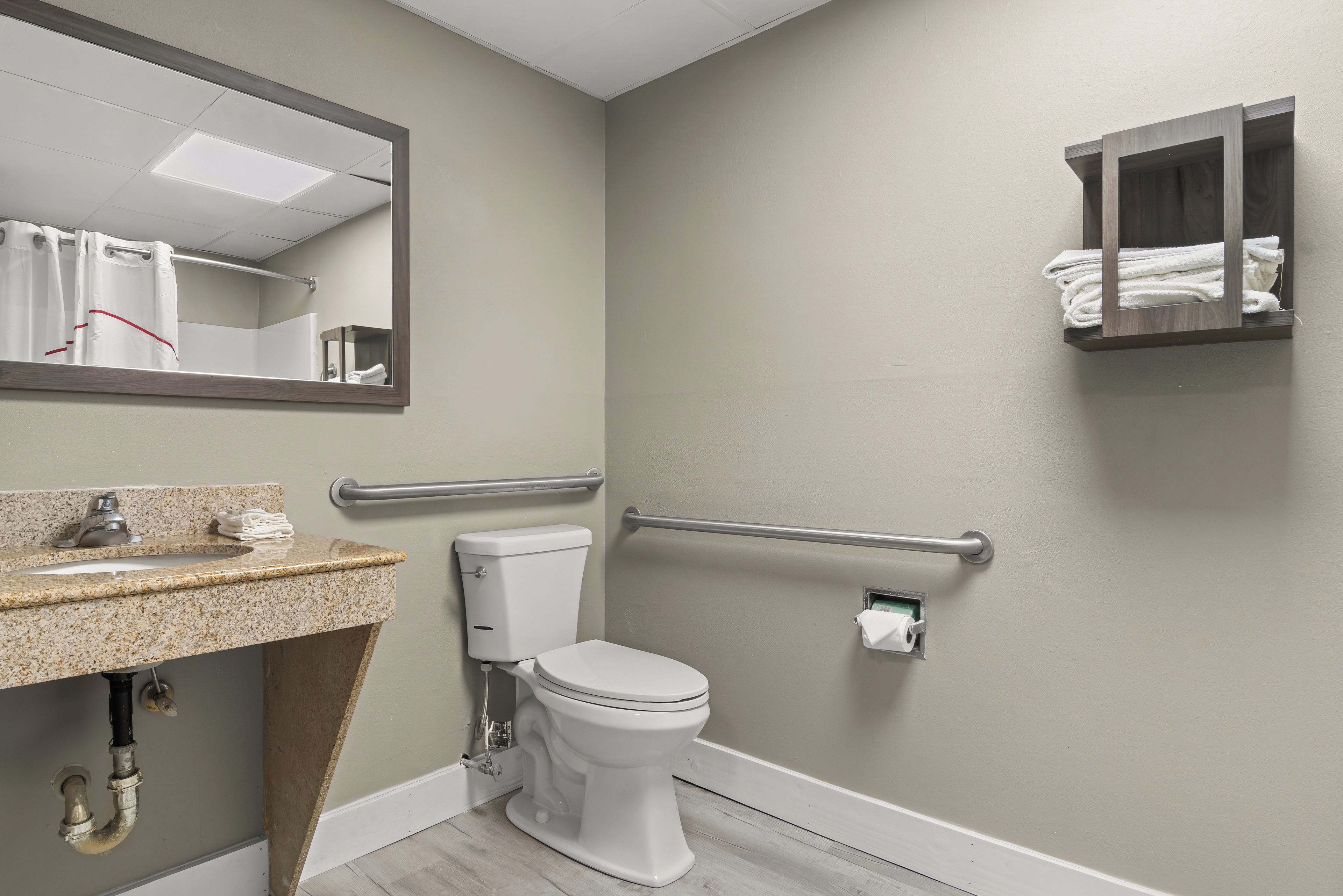 A bathroom with a toilet, sink, mirror, towel rack, and handrails.