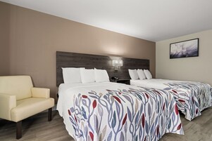 Habitación de hotel con dos camas, una silla, una mesita de noche, una lámpara y un cuadro en la pared.