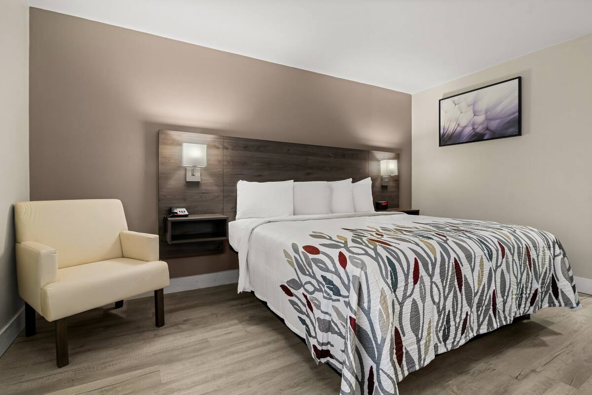 A hotel room with a bed, a chair, a nightstand, and a wall-mounted lamp.