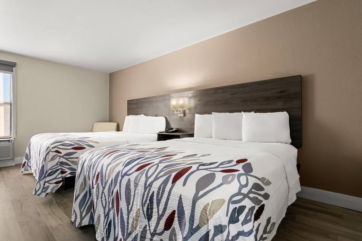A hotel room with two beds, a nightstand, a wall-mounted lamp, and a window.