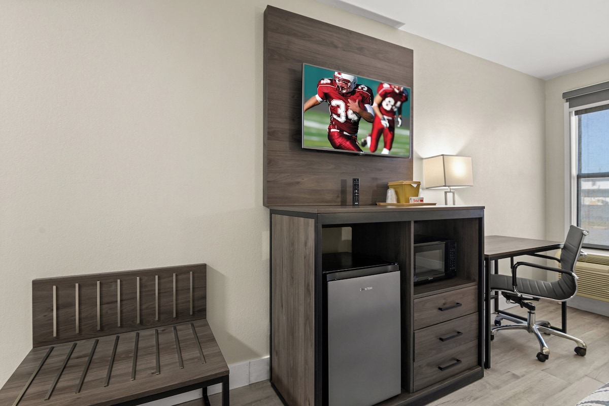 A hotel room with a wooden bench, a desk with a computer, a refrigerator, a TV mounted on the wall, and a window with a view.