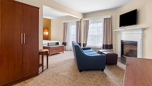 A hotel room with a large wooden wardrobe, a blue sofa, a small table, a bed, and a fireplace. - Comfort Suites near Camp Lejeune (Jacksonville)