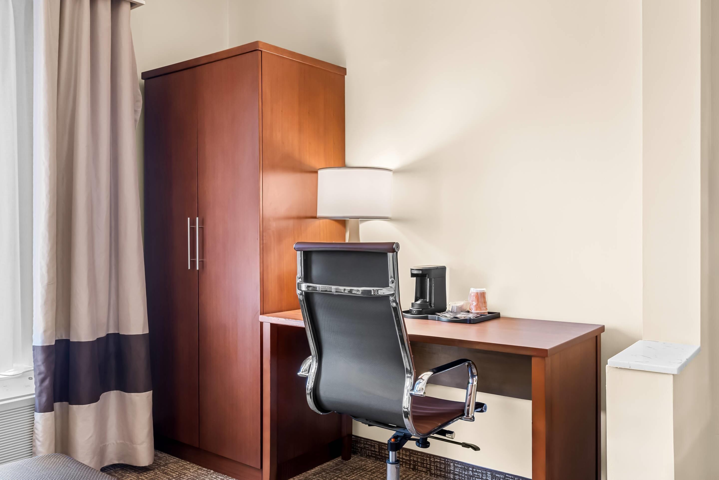 A hotel room with a wooden wardrobe, a desk with a lamp, and a chair.