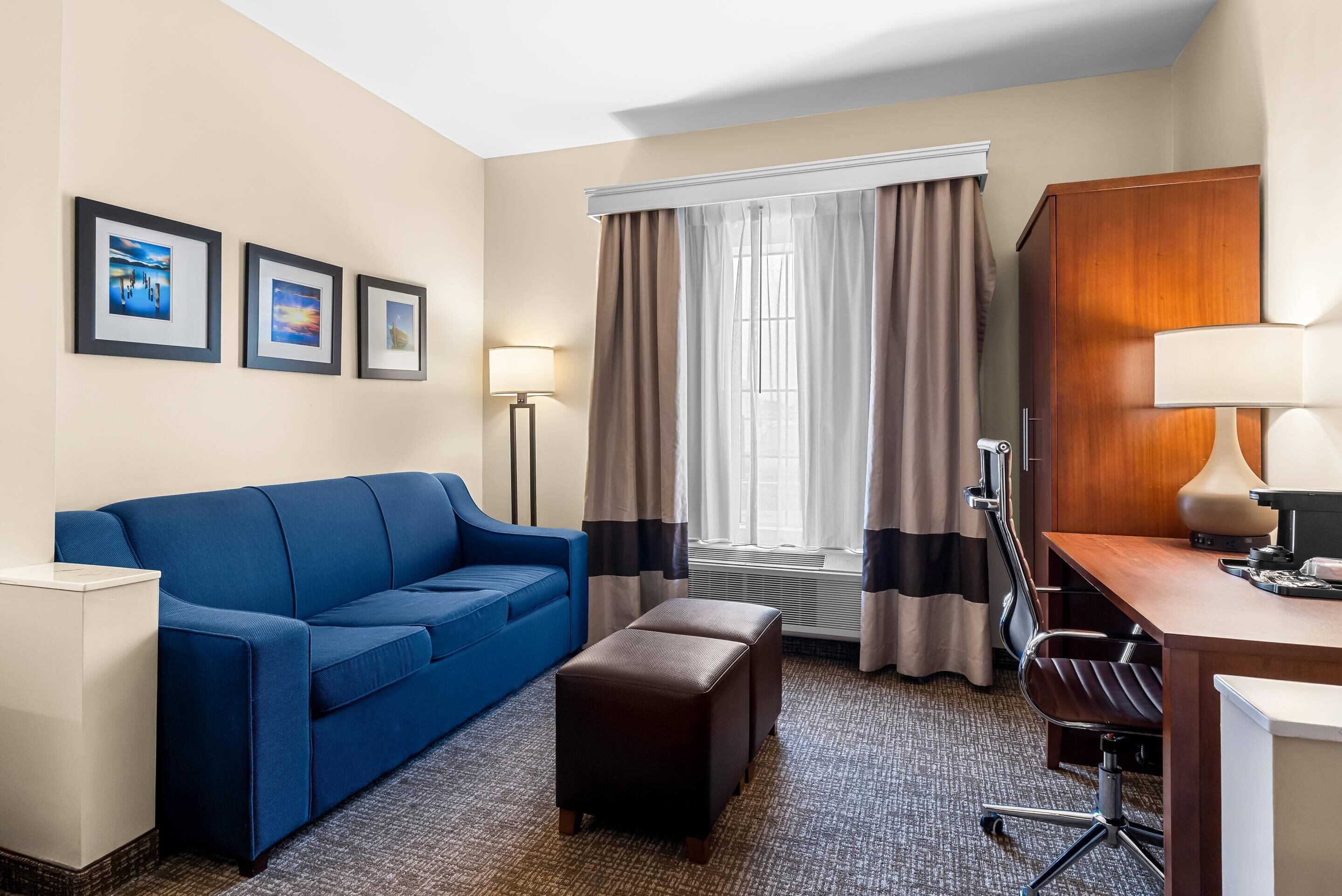 A hotel room with a blue sofa, a wooden desk, a chair, a small ottoman, and a window with curtains.
