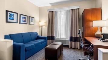 A hotel room with a blue sofa, a wooden desk, a chair, a small ottoman, and a window with curtains.