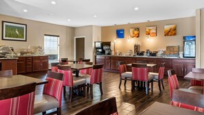 A modern dining area with red and grey upholstered chairs, a kitchen area with a coffee machine, and artwork on the walls. - Comfort Suites near Camp Lejeune (Jacksonville)