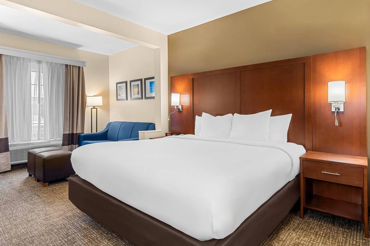 Ein Hotelzimmer mit einem großen Bett, einem Nachttisch, einer Lampe, einem Sessel und einem Fenster mit Vorhängen.