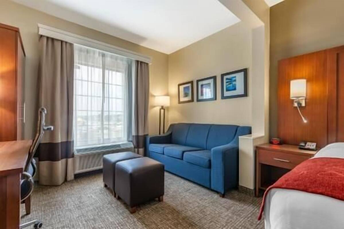 Ein Hotelzimmer mit einem Bett, einem Sofa, einem Hocker, einem Schreibtisch, einer Lampe und einem Fenster mit Vorhängen.