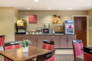Habitación de hotel con zona de comedor que incluye una mesa de madera, sillas tapizadas en rojo, microondas, máquina de café, refrigerador y dispensador de jugo.