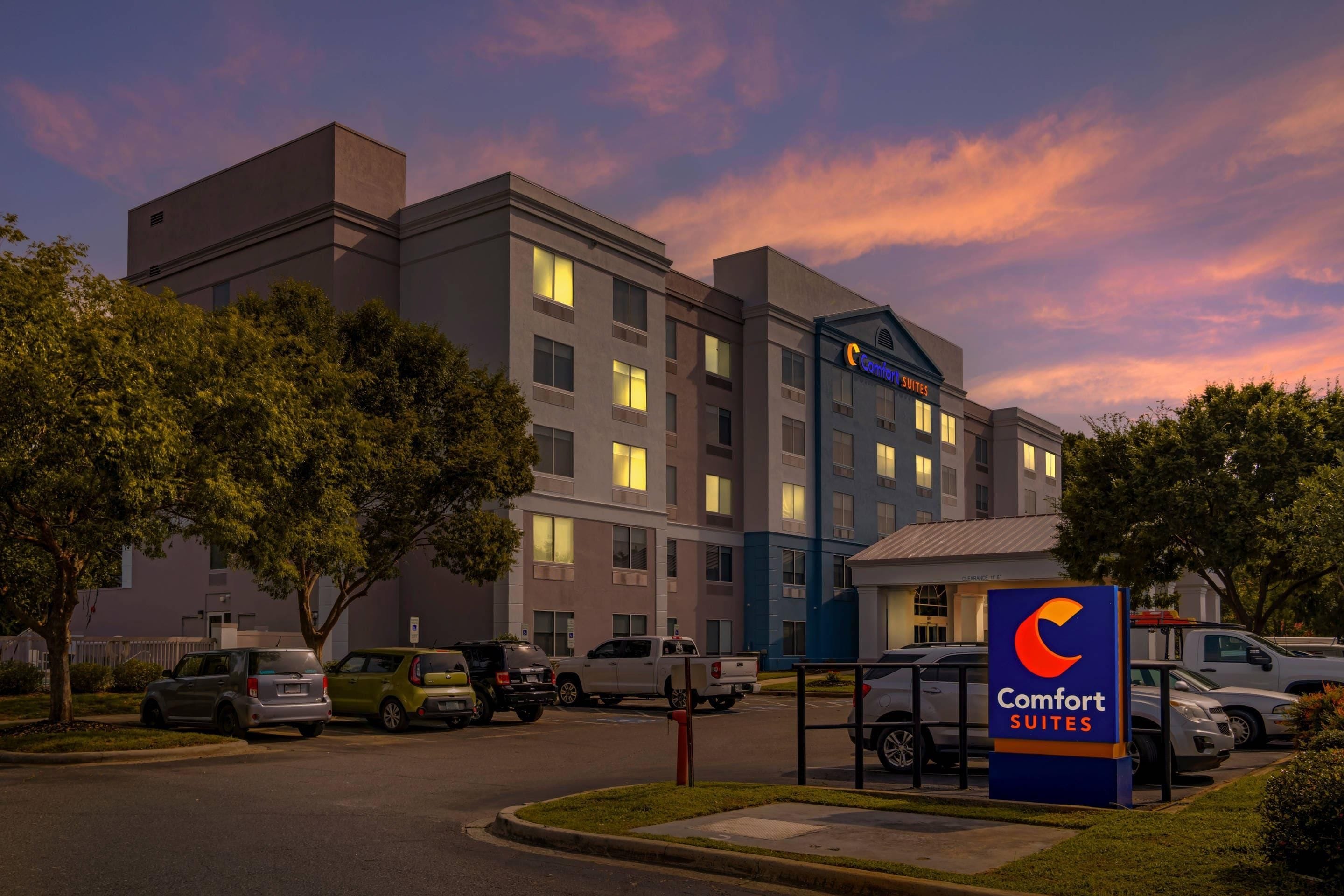 A multi-story hotel with a sign for Comfort Suites, a parking lot with various vehicles, and a clear sky.