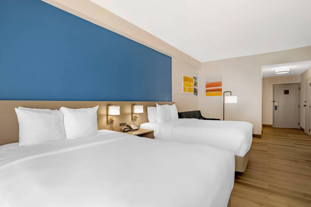 Une chambre d’hôtel avec deux lits, un mur d’accent bleu, une table de chevet avec une lampe et une porte donnant sur une autre pièce.