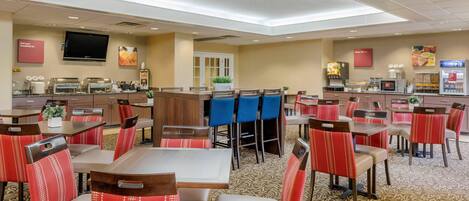 Comedor de hotel con varias mesas y sillas, zona de cocina y televisor montado en la pared.