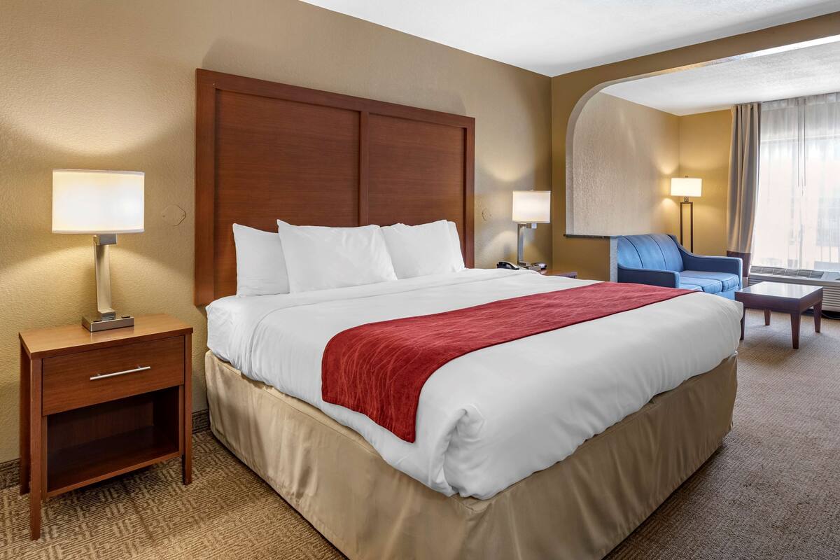 A hotel room with a large bed, a wooden nightstand, a desk, a chair, and a lamp.