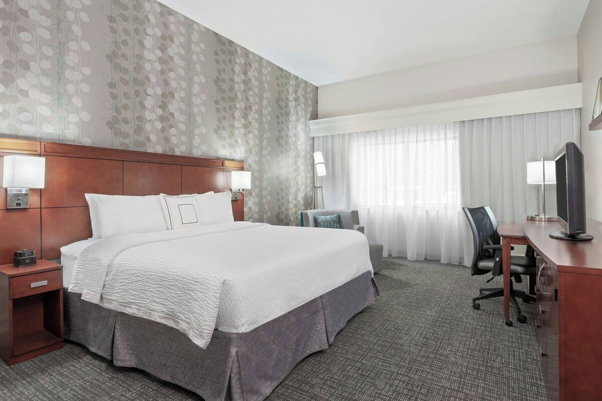 Ein Hotelzimmer mit einem großen Bett, Nachttischen, einem Schreibtisch mit Stuhl, einem Fernseher und einem Fenster mit Vorhängen.