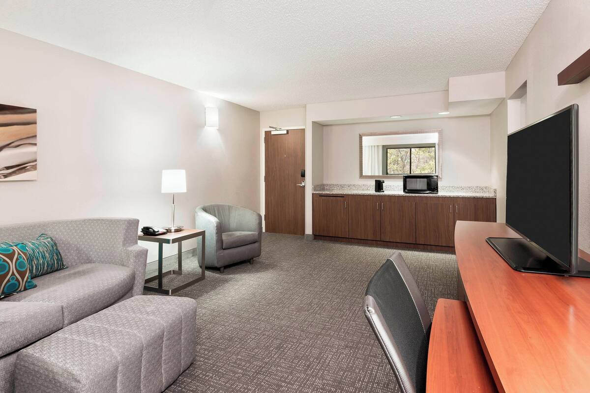 Ein modernes Hotelzimmer mit grauem Sofa, einem kleinen Sessel, einem Holztisch und einem Flachbildfernseher sowie einer Küchenzeile.