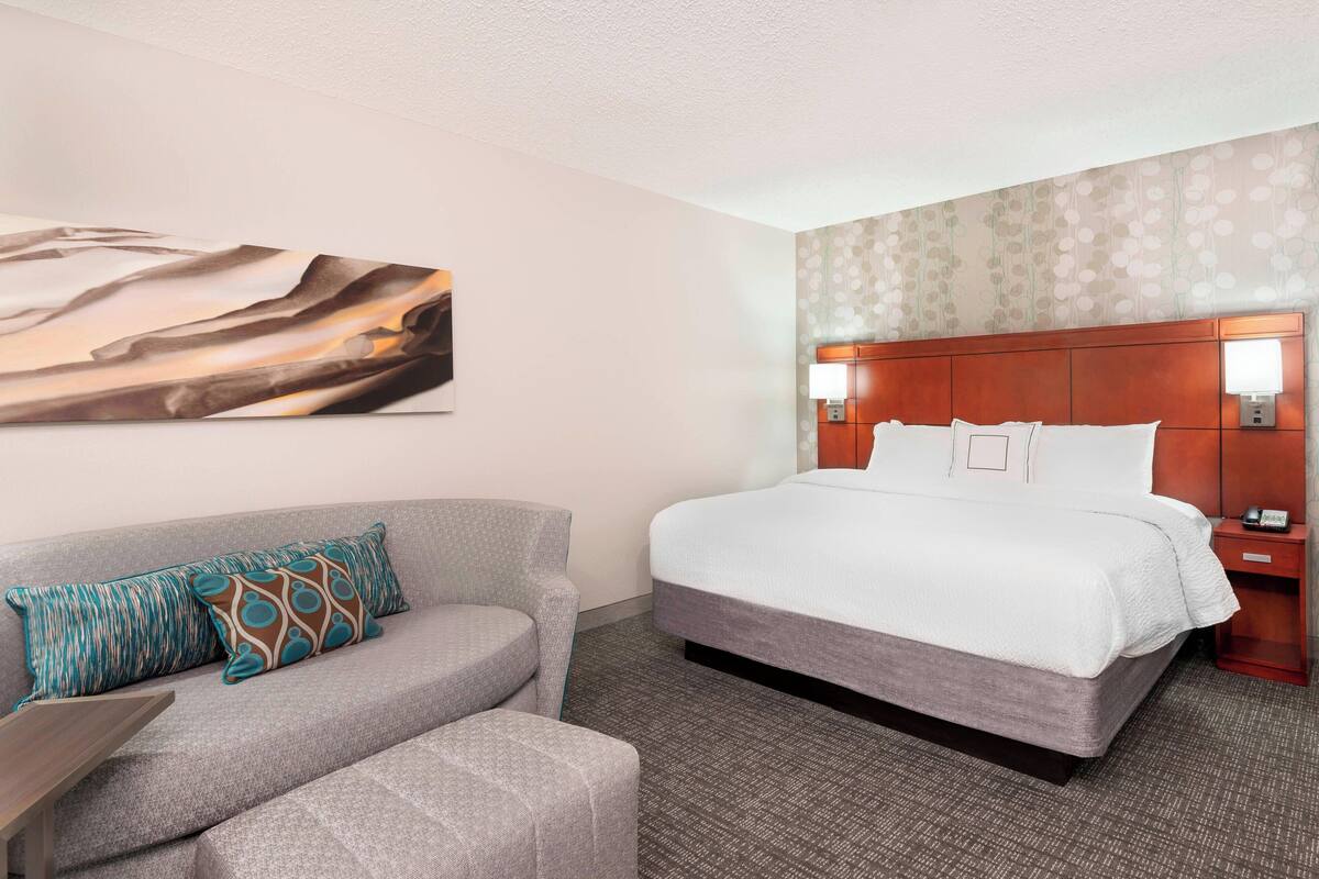 Ein Hotelzimmer mit einem großen Bett, einem grauen Sofa mit gemusterten Kissen, einem Wandbild und einem Teppich mit Muster.