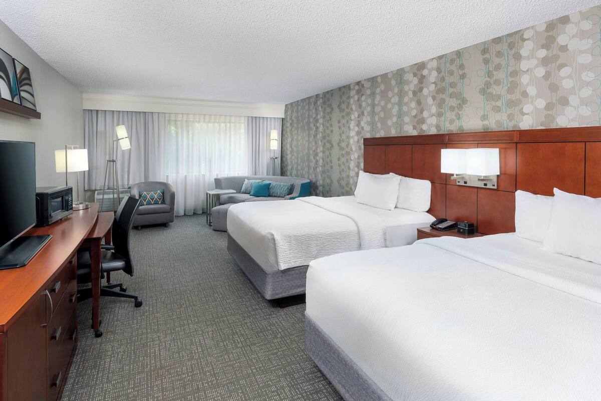 Ein Hotelzimmer mit zwei Betten, einem Schreibtisch mit Computer, einem Stuhl, einer Lampe und einem Fernseher.