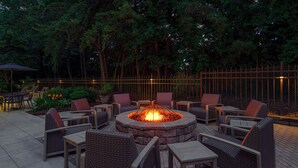 An outdoor seating area with a fire pit surrounded by wicker chairs and tables, set against a backdrop of trees and a metal fence.