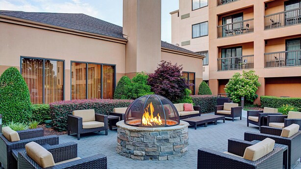An outdoor patio with a stone fire pit surrounded by wicker furniture and a large window overlooking the building. - Courtyard Durham Near Duke University/Downtown (Durham)