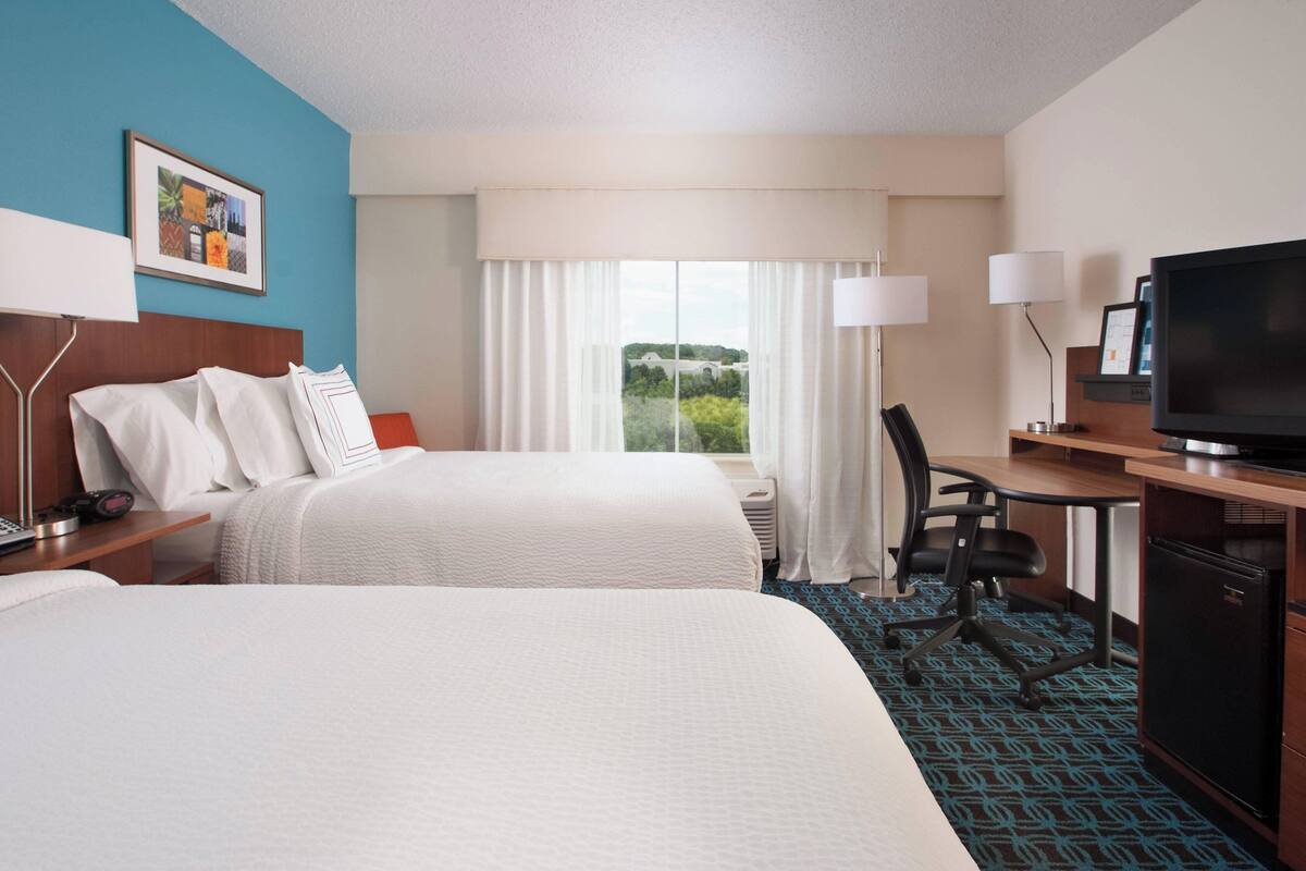 A hotel room with two beds, a desk with a television, and a large window with curtains.